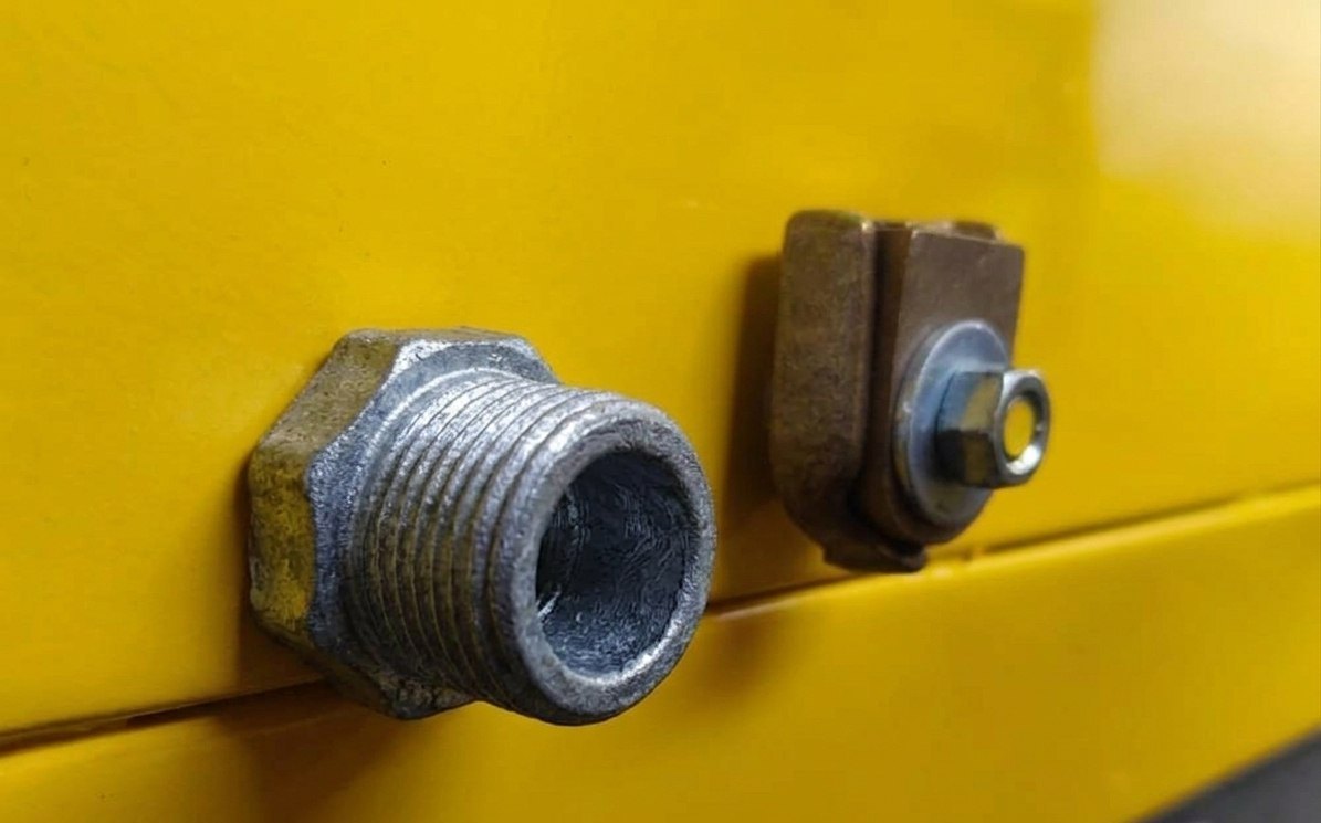 Primer plano de la pared lateral de la cuba: a la izquierda, la boca roscada de drenaje en acero galvanizado; a la derecha, el borne de bronce para la puesta a tierra, con su tuerca.